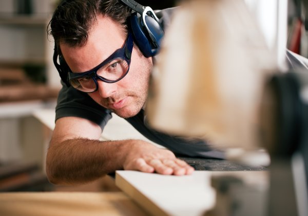 Ein Mann im schwarzen T-Shirt mit schwarzer Brille und Kopfhörern als Lärmschutz sägt ein Holzbrett an einer Maschine.