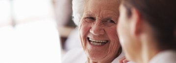 Das Bild zeigt eine lächelnde alte Frau im Gespräch mit einer anderen Person.